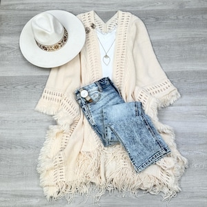 May include: A cream-colored crochet cardigan with fringe detailing, paired with light blue denim jeans and a white hat with a brown band.