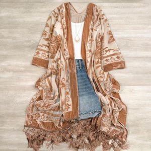 May include: A sheer, long-sleeved kimono featuring a tan and cream floral pattern with velvet-textured trim. The garment is layered over a white tank top and blue denim shorts. A gold pendant necklace hangs from the neckline, and the hem is finished with long, matching tan fringe.