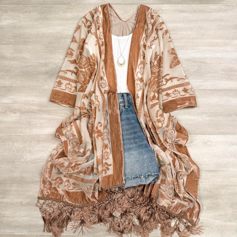 May include: A sheer, long-sleeved kimono featuring a tan and cream floral pattern with velvet-textured trim. The garment is layered over a white tank top and blue denim shorts. A gold pendant necklace hangs from the neckline, and the hem is finished with long, matching tan fringe.