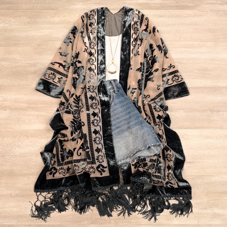 May include: A beige kimono featuring a dark charcoal velvet floral pattern, a long fringe hem, and wide sleeves. The garment is layered over a white tank top with a gold pendant necklace and blue denim shorts with a raw, frayed hemline.