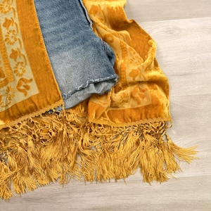 May include: A mustard yellow velvet scarf featuring a floral pattern, decorative braided trim, and long fringe tassels, layered over a pair of blue denim shorts with a frayed hem. The fabric has a soft texture with visible light and dark tones.