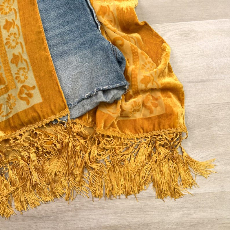 May include: A mustard yellow velvet scarf featuring a floral pattern, decorative braided trim, and long fringe tassels, layered over a pair of blue denim shorts with a frayed hem. The fabric has a soft texture with visible light and dark tones.