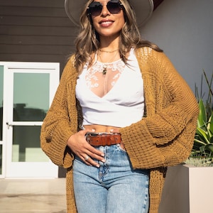 May include: A woman wearing a white hat, sunglasses, a white top, a brown crocheted cardigan, and blue jeans with a brown belt.