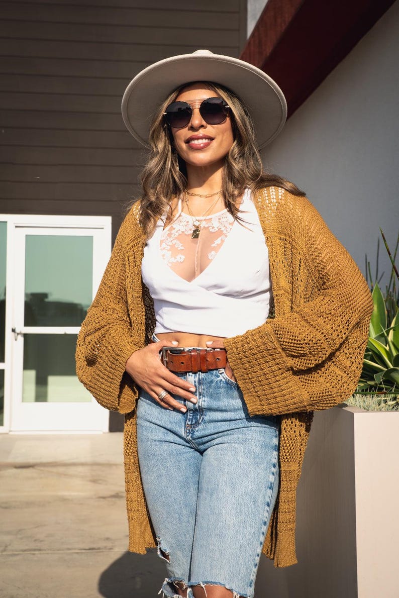 May include: A woman wearing a white hat, sunglasses, a white top, a brown crocheted cardigan, and blue jeans with a brown belt.
