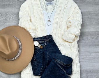 May include: An ivory cable-knit cardigan, white tank top, and dark wash denim jeans are arranged with a brown hat, silver necklace, and a wristwatch. The hat has a decorative band. The outfit is styled on a light gray wood-grain surface.
