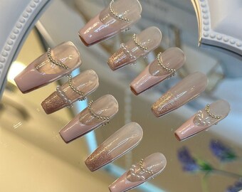 May include: A set of ten false nails with a nude base and a glitter accent. Each nail is decorated with a pearl and gold chain.