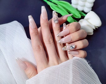 May include: Close-up of a hand with long, clear, oval-shaped fake nails with silver tips and rhinestones. The nails are decorated with small, clear rhinestones and gold accents.