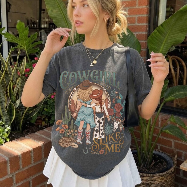 May include: A charcoal gray short-sleeve t-shirt featuring a graphic print of two figures in cowboy hats, cacti, flowers, and a snake. The text reads COWGIRL SLIKE & ME in a decorative font. The shirt is paired with a white pleated skirt.