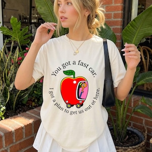 Könnte beinhalten: Ein cremefarbenes Kurzarm-T-Shirt mit einer Grafik eines roten Apfels in Form eines Autos mit einem Wurm darin. Der Text "You got a fast car. I got a plan to get us out of here." ist in einer kreisförmigen schwarzen Schrift um die Grafik gedruckt.