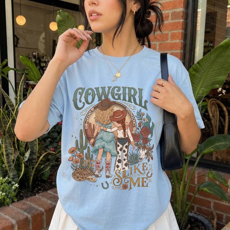 May include: A light blue t-shirt featuring a graphic print of two figures wearing cowboy hats and boots, surrounded by cacti, flowers, and a skull. The text "COWGIRL SLIKE ME" is printed in a decorative font above and below the illustration.