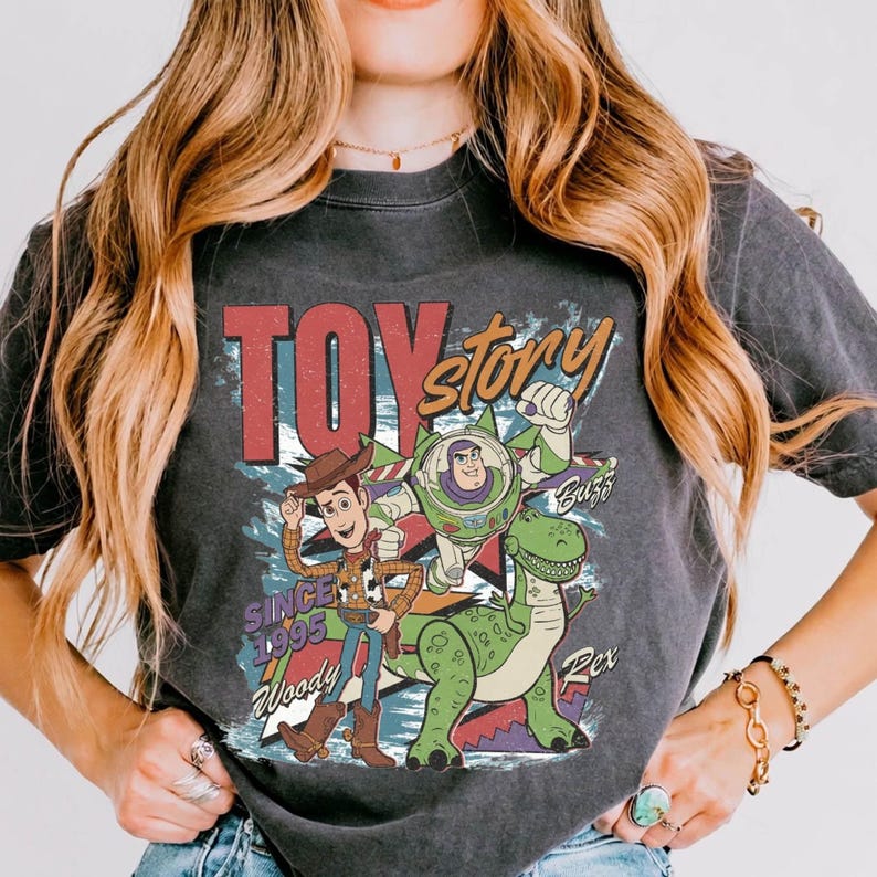 May include: A charcoal gray t-shirt featuring a vintage-style graphic of Woody, Buzz Lightyear, and Rex from Toy Story. The design includes the text "TOY story", "SINCE 1995", "Woody", "Buzz", and "Rex" in a distressed, colorful print on the front.