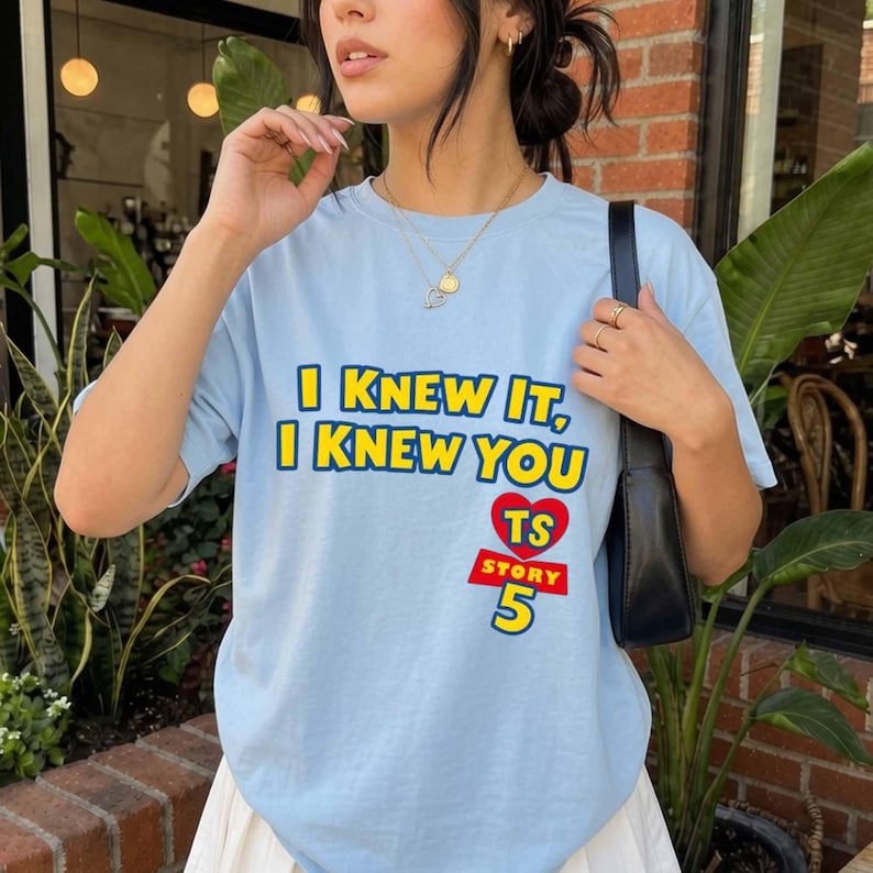 Może przedstawiać: Jasnoniebieski t-shirt z kr&oacute;tkim rękawem z ż&oacute;łtym napisem I KNEW IT, I KNEW YOU nad czerwonym sercem z literami TS, słowem STORY w kolorze białym na czerwonym prostokącie oraz ż&oacute;łtą cyfrą 5 pod sercem.