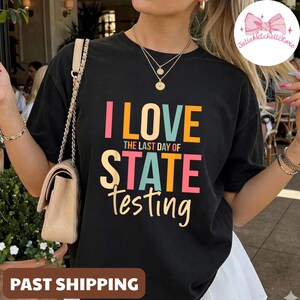 Può includere: T-shirt nero con la scritta "I LOVE THE LAST DAY OF STATE TESTING" in rosa, arancione, turchese e giallo. La maglietta ha una vestibilità ampia ed è indossata da una persona. È visibile una piccola borsa con tracolla a catena.