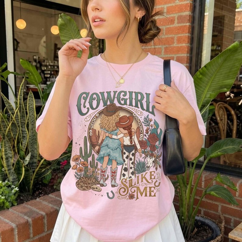 May include: A light pink short-sleeve t-shirt featuring a graphic print of two figures in cowboy hats, cacti, flowers, and a snake. The text reads COWGIRL SLIKE ME in a decorative font. The shirt is worn with a black shoulder bag and gold necklaces.