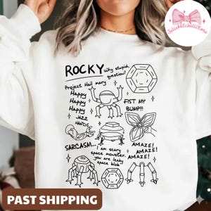 Könnte beinhalten: Weißes Sweatshirt mit einem schwarzen Grafikdesign. Das Design enthält das Wort "ROCKY", Cartoon-Illustrationen und Sätze wie "Project Hail Mary", "Happy", "Jazz Hands" und "Sarcasm".
