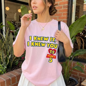 Może przedstawiać: Jasnor&oacute;żowy t-shirt z kr&oacute;tkim rękawem z ż&oacute;łtym napisem I KNEW IT, I KNEW YOU, czerwonym sercem z literami TS oraz czerwonym napisem STORY 5. Koszulka jest noszona z czarną torbą na ramię i złotym naszyjnikiem z małym wisiorkiem.