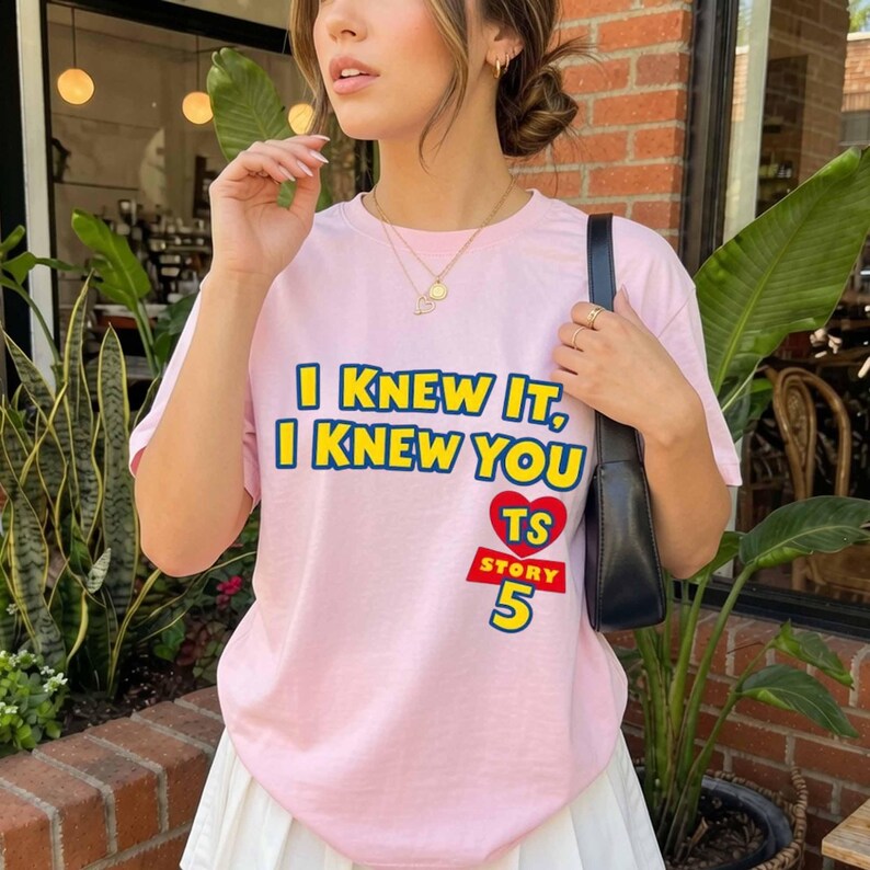 Może przedstawiać: Jasnor&oacute;żowy t-shirt z kr&oacute;tkim rękawem z ż&oacute;łtym napisem I KNEW IT, I KNEW YOU, czerwonym sercem z literami TS oraz czerwonym napisem STORY 5. Koszulka jest noszona z czarną torbą na ramię i złotym naszyjnikiem z małym wisiorkiem.