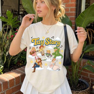 May include: A cream-colored short-sleeve t-shirt featuring the text Tay Story in yellow letters. The graphic includes six illustrated characters on white clouds, surrounded by musical notes and yellow stars, with a black shoulder bag strap.