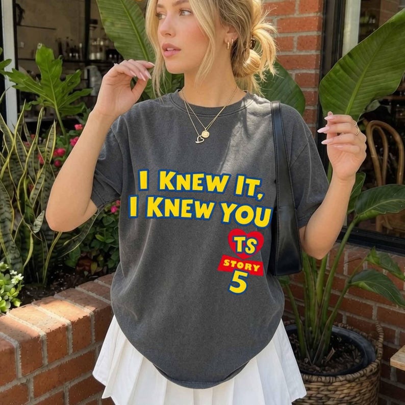 Może przedstawiać: Ciemnoszary t-shirt z kr&oacute;tkim rękawem z ż&oacute;łtym napisem I KNEW IT, I KNEW YOU, czerwoną grafiką serca z literami TS oraz słowem STORY zapisanym białymi literami nad dużą ż&oacute;łtą cyfrą 5 z przodu ubrania.