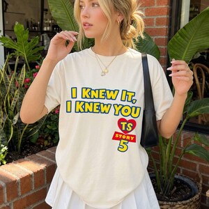 Może przedstawiać: Kremowy t-shirt z kr&oacute;tkim rękawem z niebieskim napisem I KNEW IT, I KNEW YOU, czerwonym sercem z literami TS oraz czerwonym napisem STORY 5 poniżej. Koszulka ma okrągły dekolt i luźny kr&oacute;j, noszona z czarną torebką na ramię.