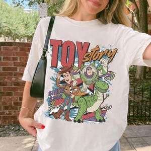 May include: A white short-sleeve t-shirt featuring a graphic print of Woody, Buzz Lightyear, and Rex from Toy Story. The design includes the text Toy Story, Since 1995, Woody, Buzz, and Rex in colorful lettering against a blue and red background.