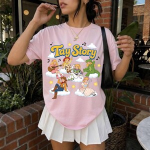 May include: A light pink short-sleeve t-shirt featuring the text Tay Story in yellow and blue letters. The graphic includes six illustrated characters sitting on white clouds, surrounded by yellow stars and black musical notes.