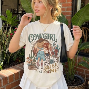 May include: A cream-colored short-sleeve t-shirt featuring a graphic print of two figures in cowboy hats with the text COWGIRL SLIKE ME. The illustration includes desert elements like cacti, flowers, and a snake, with a western-themed color palette.