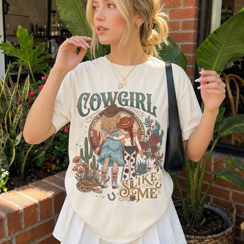 May include: A cream-colored short-sleeve t-shirt featuring a graphic print of two figures in cowboy hats with the text COWGIRL SLIKE ME. The illustration includes desert elements like cacti, flowers, and a snake, with a western-themed color palette.