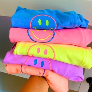 Op de afbeelding: Een stapel opgevouwen sweatshirts in heldere kleuren: blauw, roze, geel, paars en oranje. Elke sweatshirt heeft een groot smiley-gezichtontwerp met een contrasterende omtrek en oogkleur.
