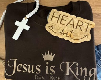 May include: A brown sweatshirt with the text "Jesus is King" and "REV 19:16" printed on it. A gold crown is above the text. A wooden sign with the text "HEART set" is on top of the sweatshirt.