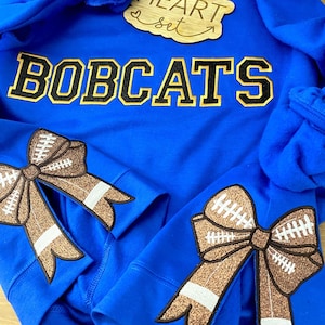 May include: Royal blue sweatshirt with the word "BOBCATS" in black and gold letters. The sleeves have brown and white football-themed bows.