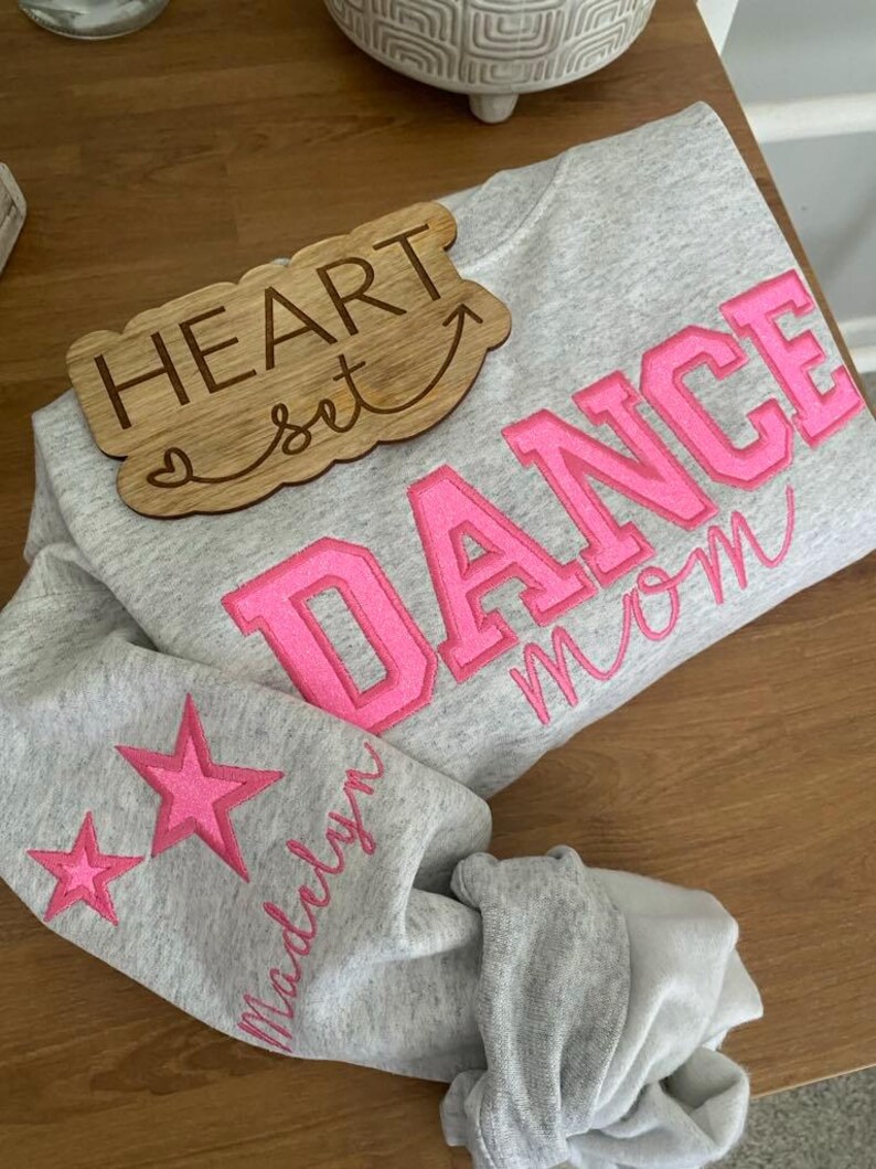 May include: A gray sweatshirt with the words "DANCE MOM" in bright pink block letters. The sleeve features two pink stars and the name "Madelyn" in pink script. A wooden sign that says "HEART set" is also in the image.