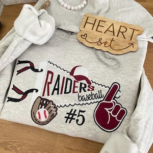 May include: A gray hooded sweatshirt with a red and white baseball themed design. The design includes a baseball glove, a baseball, a pennant with the word "RAIDERS" and the text "baseball", and a pointing finger. The number "#5" is also included in the design. The sweatshirt has a wooden sign attached to the hood that says "HEART set".