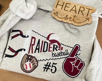 May include: A gray hooded sweatshirt with a red and white baseball themed design. The design includes a baseball glove, a baseball, a pennant with the word "RAIDERS" and the text "baseball", and a pointing finger. The number "#5" is also included in the design. The sweatshirt has a wooden sign attached to the hood that says "HEART set".
