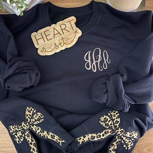 May include: Black sweatshirt with leopard print bows on the sleeves and a monogrammed "HH" on the front. A wooden sign with the text "HEART set" is placed on top of the sweatshirt.