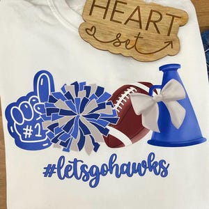 May include: White t-shirt with blue and gray graphics of a foam finger, pom-pom, football, and megaphone. The text "#letsgohawks" is below the graphics. A wooden heart-shaped sign that says "HEART set" is on top of the shirt.