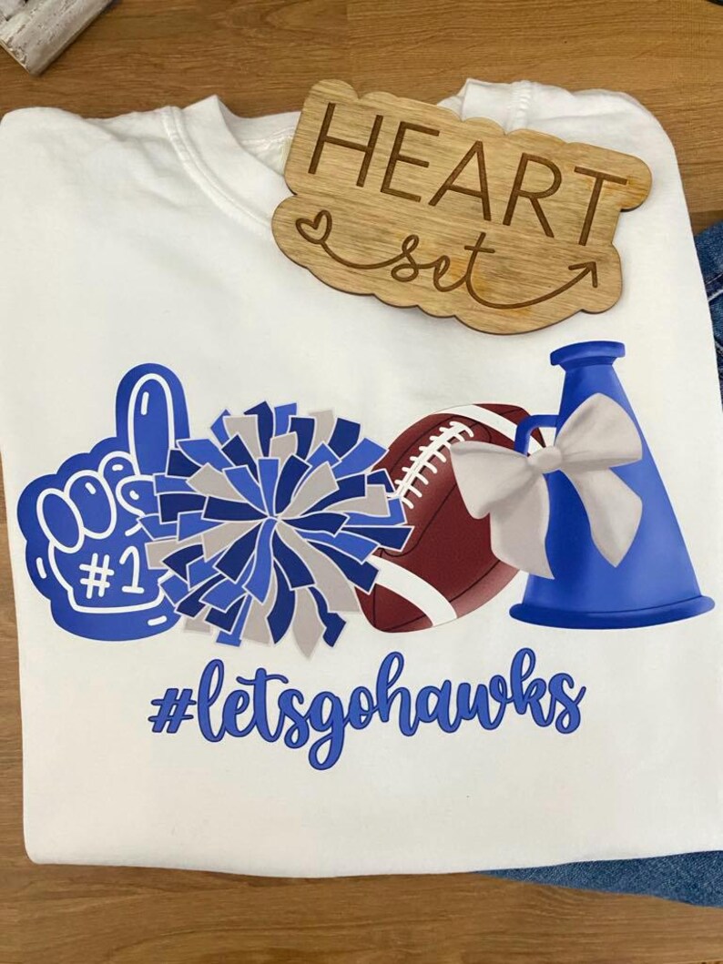 May include: White t-shirt with blue and gray graphics of a foam finger, pom-pom, football, and megaphone. The text "#letsgohawks" is below the graphics. A wooden heart-shaped sign that says "HEART set" is on top of the shirt.