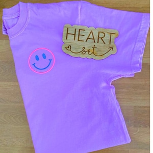 Op de afbeelding: Een lila t-shirt met een smiley-gezicht in een roze cirkel en blauwe details. Een houten bord met de woorden "HEART set" en een pijl is op het shirt geplaatst. Het shirt ligt op een houten oppervlak.