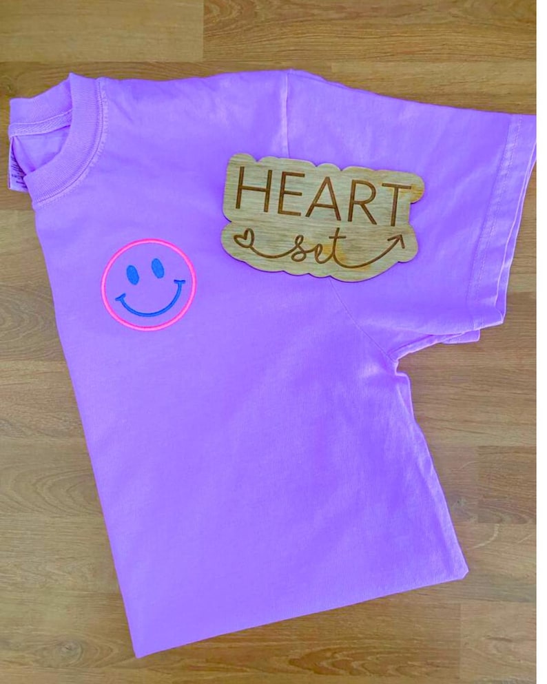Op de afbeelding: Een lila t-shirt met een smiley-gezicht in een roze cirkel en blauwe details. Een houten bord met de woorden "HEART set" en een pijl is op het shirt geplaatst. Het shirt ligt op een houten oppervlak.