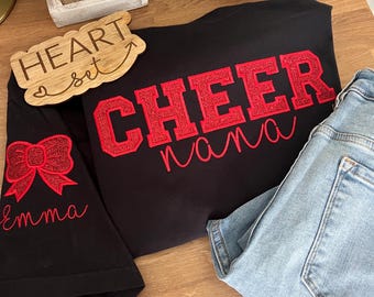 May include: Black t-shirt with red glitter lettering that says "Cheer Nana" and a red glitter bow on the sleeve. The text "Emma" is written in white on the sleeve.