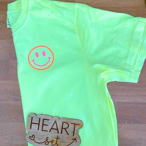 Op de afbeelding: Een neongeel t-shirt met een ronde smiley-gezicht afbeelding op de linkerborst. De smiley heeft een roze centrum en een oranje omtrek. Een houten bord met de woorden "HEART set" is op het shirt geplaatst.