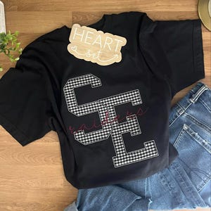 May include: Black t-shirt with a gingham-patterned "SE" design and the word "Raiders" in cursive. A wooden sign that says "HEART set" is on top of the shirt. The shirt is paired with blue jeans.