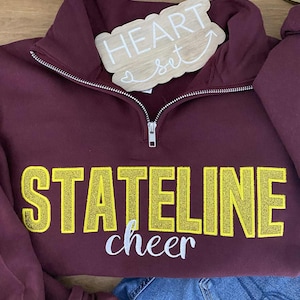 May include: A maroon quarter-zip sweatshirt with the words "STATELINE cheer" in yellow and white lettering. A small wooden sign that says "HEART set" is on top of the sweatshirt. The sweatshirt is paired with blue jeans.