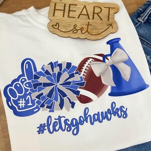 May include: White crew neck t-shirt with a blue and gray pom-pom, football, and megaphone graphic. The shirt also features a blue #1 finger, the text "#letsgohawks", and a wooden sign that says "HEART set".