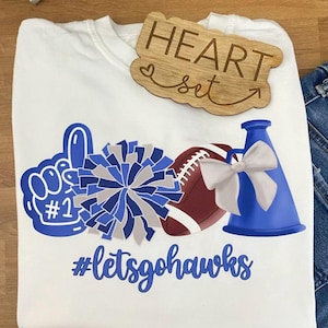 May include: White t-shirt with a blue and white graphic design featuring a foam finger, pom-pom, football, and megaphone. The text "#letsgohawks" is below the graphics. A wooden sign that says "HEART set" is on top of the shirt.