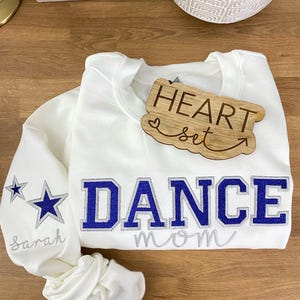 May include: White sweatshirt with the words "DANCE MOM" in blue and silver lettering. The left sleeve has two star graphics and the name "Sarah" embroidered in silver. A wooden heart-shaped tag says "HEART set".