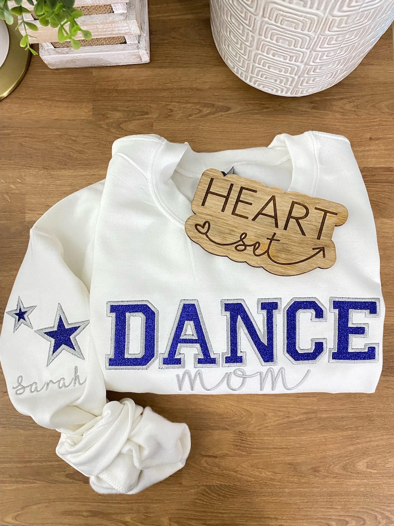May include: White sweatshirt with the words "DANCE MOM" in blue and silver lettering. The left sleeve has two star graphics and the name "Sarah" embroidered in silver. A wooden heart-shaped tag says "HEART set".