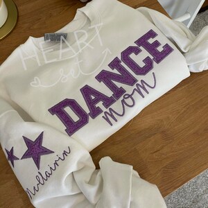 May include: White sweatshirt with the words "HEART set DANCE mom" in a mix of script and block letters. The word "DANCE" is in purple glitter. The left sleeve has two purple glitter stars and the name "Mcllairin" in script.