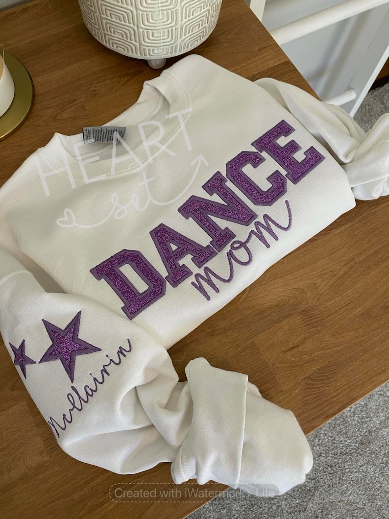 May include: White sweatshirt with the words "HEART set DANCE mom" in a mix of script and block letters. The word "DANCE" is in purple glitter. The left sleeve has two purple glitter stars and the name "Mcllairin" in script.