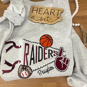 May include: Gray sweatshirt with a maroon and black pennant that says "RAIDERS" with a basketball, baseball glove, and a maroon hand with a finger pointing up. The text "Brayden" is below the pennant. A wooden sign that says "HEART set" is above the pennant.